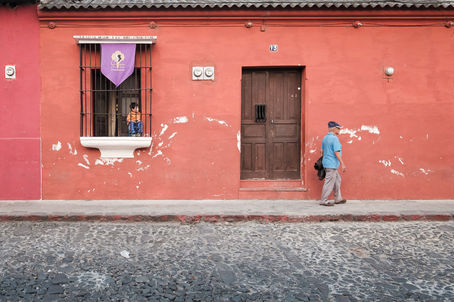 A young boy watches an elder closely from the window of his home. Under the symbolic purple banner that adorns his window, his eyes are wide and perceptive of the festivities beginning to unfold in front of him.