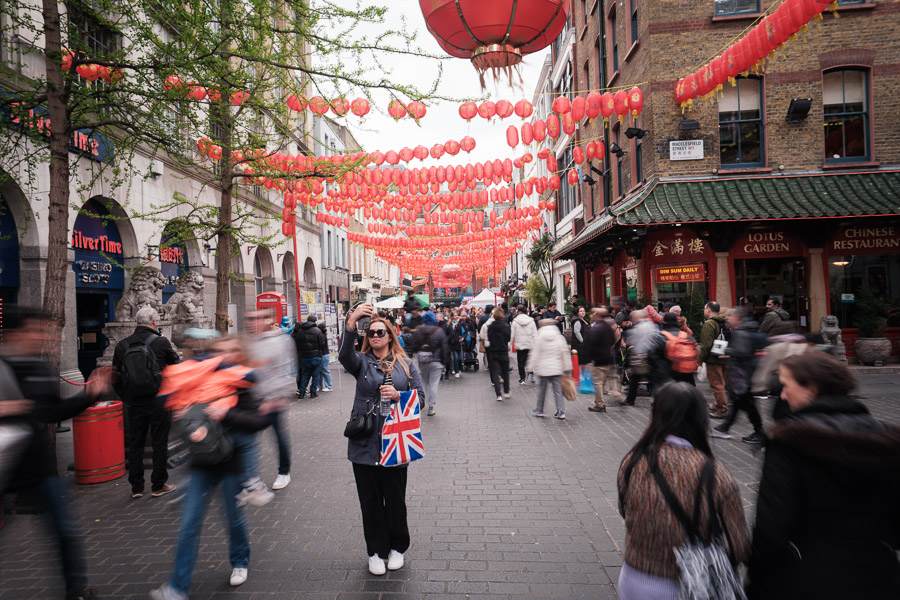 A woman with a Union Jack tote takes a selfie beneath strings of red lanterns in London's Chinatown as crowds blur around her