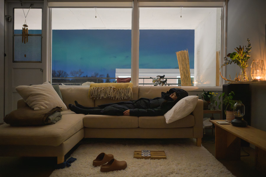 A person naps on a couch in a parka while the aurora glows teal through the apartment's living room windows